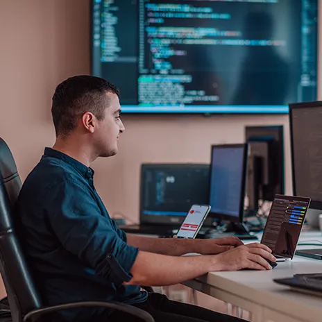 Softwareentwickler arbeitet an einem Laptop, umgeben von mehreren Bildschirmen, auf denen Code und Software-Entwicklungsumgebungen angezeigt werden.