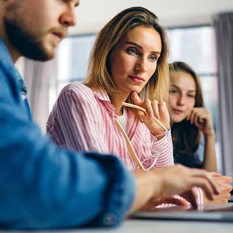 IT-Sicherheitsexperte analysiert Code auf einem Bildschirm, während im Hintergrund Kollegen über Cybersecurity-Strategien diskutieren. Mehrere Monitore mit Code- und Sicherheitsanalysen sichtbar.