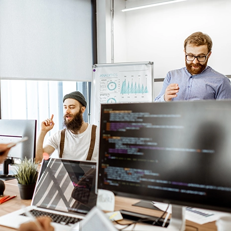 IT-Sicherheitsexperte analysiert Code auf einem Bildschirm, während im Hintergrund Kollegen über Cybersecurity-Strategien diskutieren. Mehrere Monitore mit Code- und Sicherheitsanalysen sichtbar.